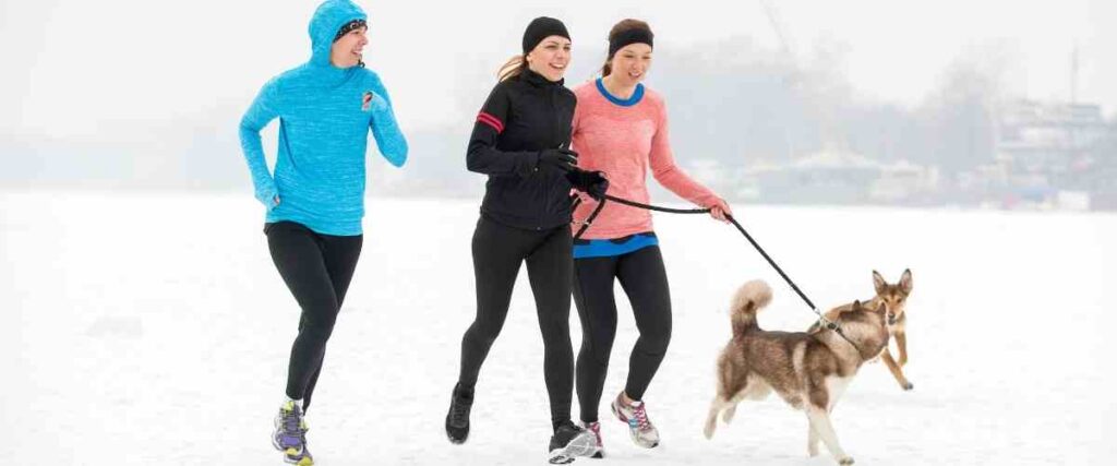 3 women running with dog on leash with another dog off leash coming up to them mid-run.