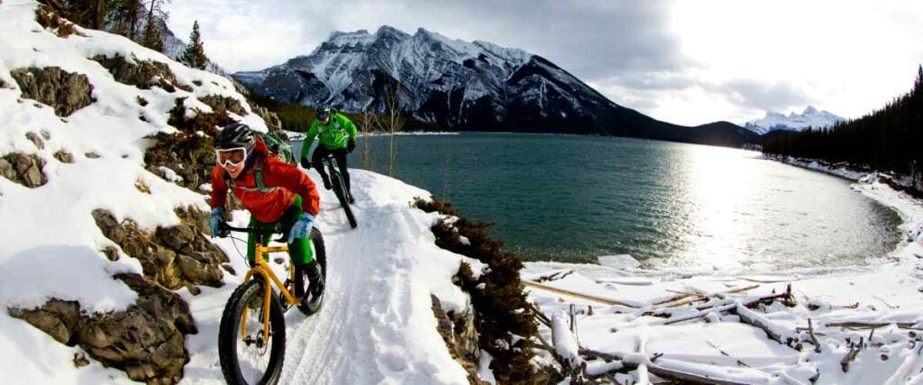 Two cyclists on a winter trail with a lake and mountains in the backyard. 