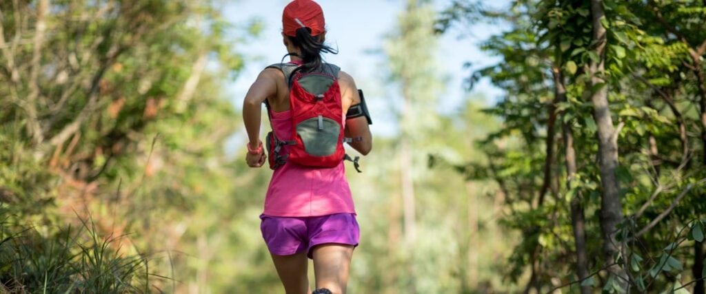 A woman on a trail with her backpack, hat and sunglasses while on a run.